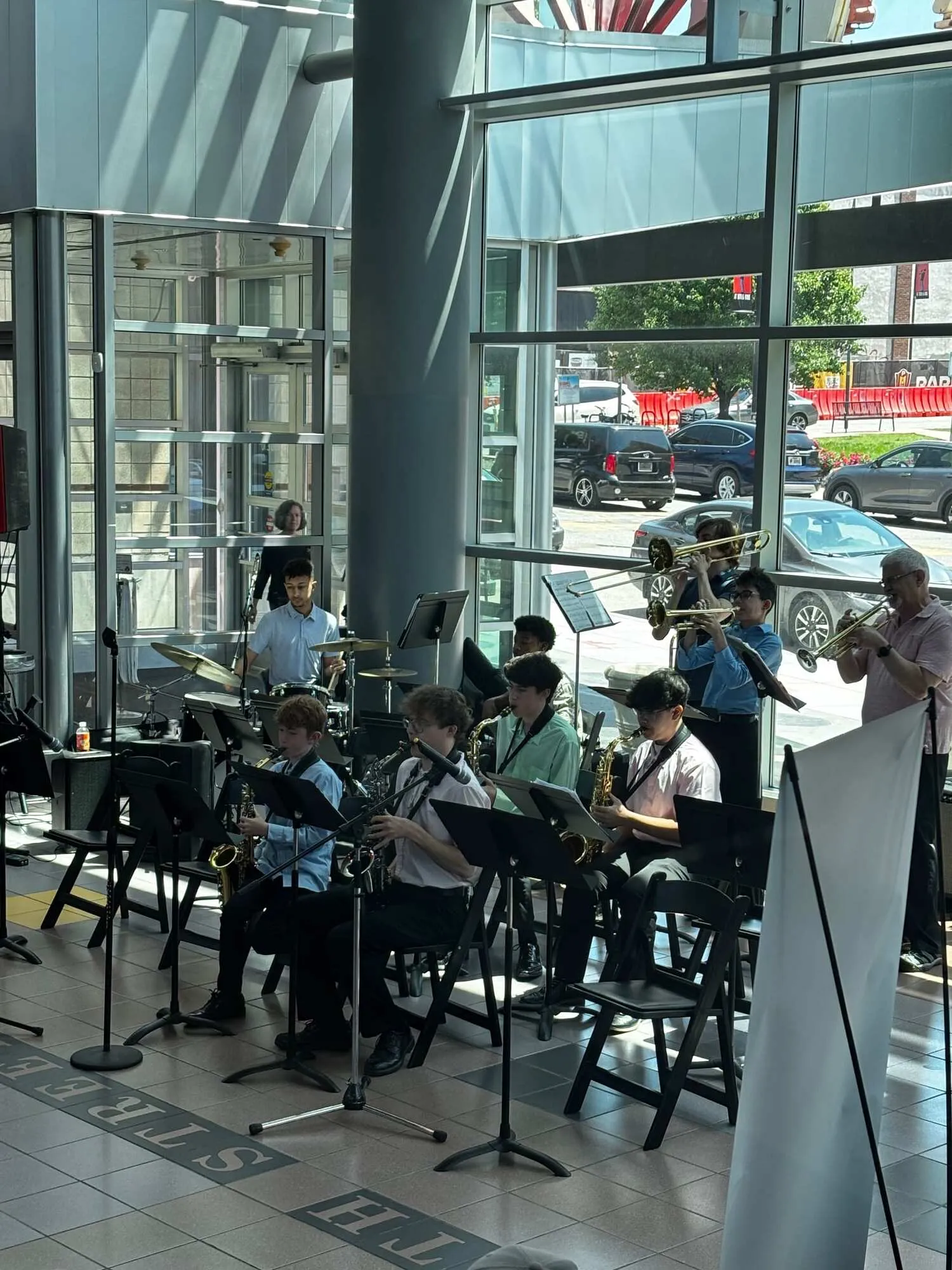 The J. MIQ Combo performs together, students playing saxophones and other instruments in a sunlit atrium.